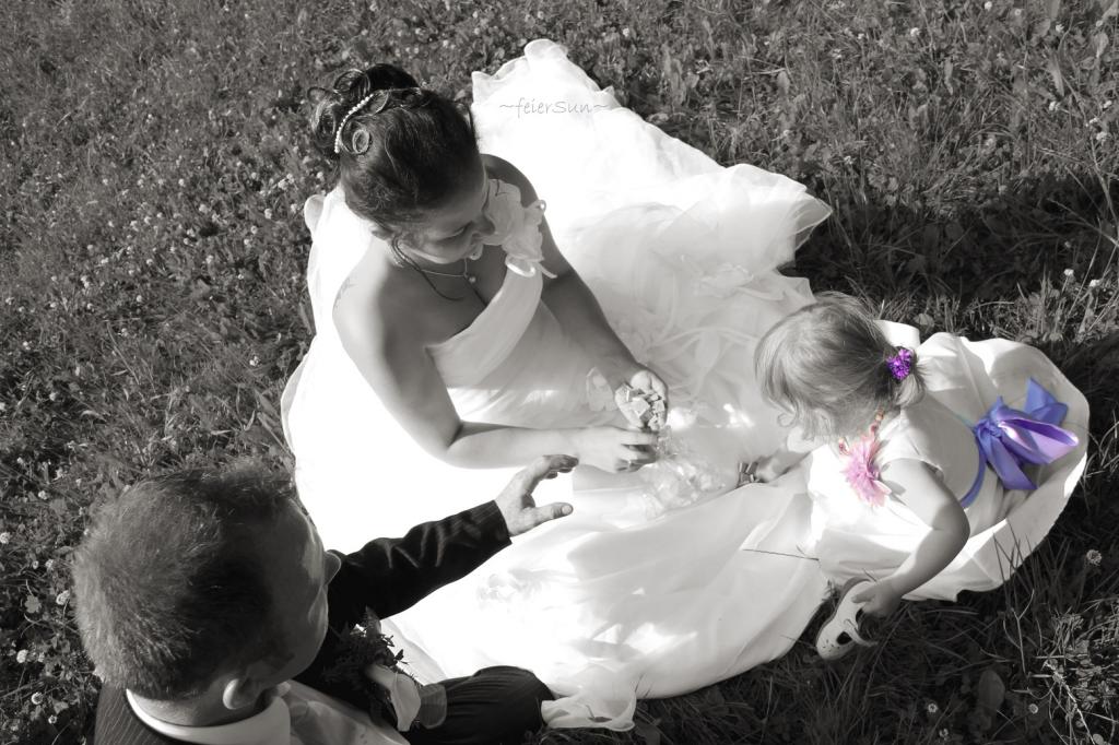 Paar mit Kind sitzt auf einer Wise und legt Buchstaben zusammen. Frau Trägt Hochzeitskleid und Mann Anzug. Das Kind ein weißes kleid mit lilafarbener Schleife. 