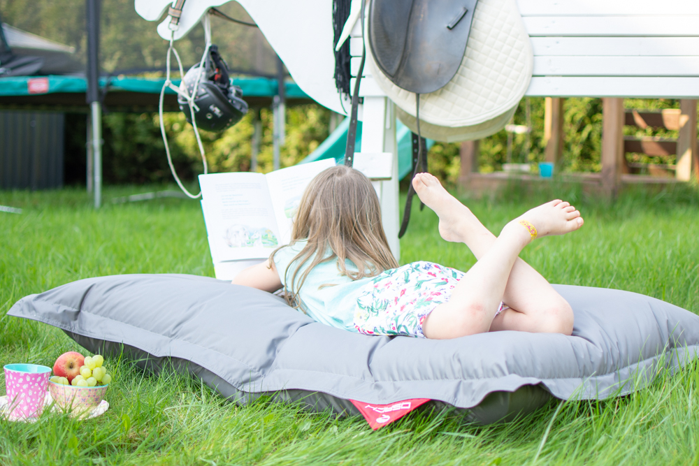 Mädchen liegt auf einem grauen Sitzsack von QSacks und liest ein Buch über Pferde und Fohlen. Auf dem Bild steht folgende Schrift "www.feiersun.de, Pause, Abenteuer und zwischen 2 Abenteuern liegt eine Pause"